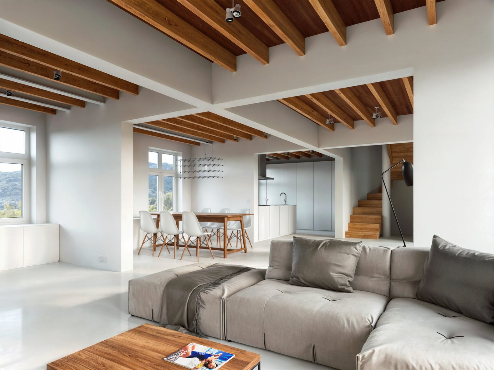 Modern open-plan living area with a spacious grey sofa, wooden ceiling beams, and a kitchen in the background.